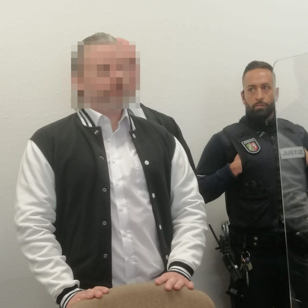 Das Bild zeigt den Angeklagten, der von einem Justizbeamten in den Gerichtssaal gebracht wird.