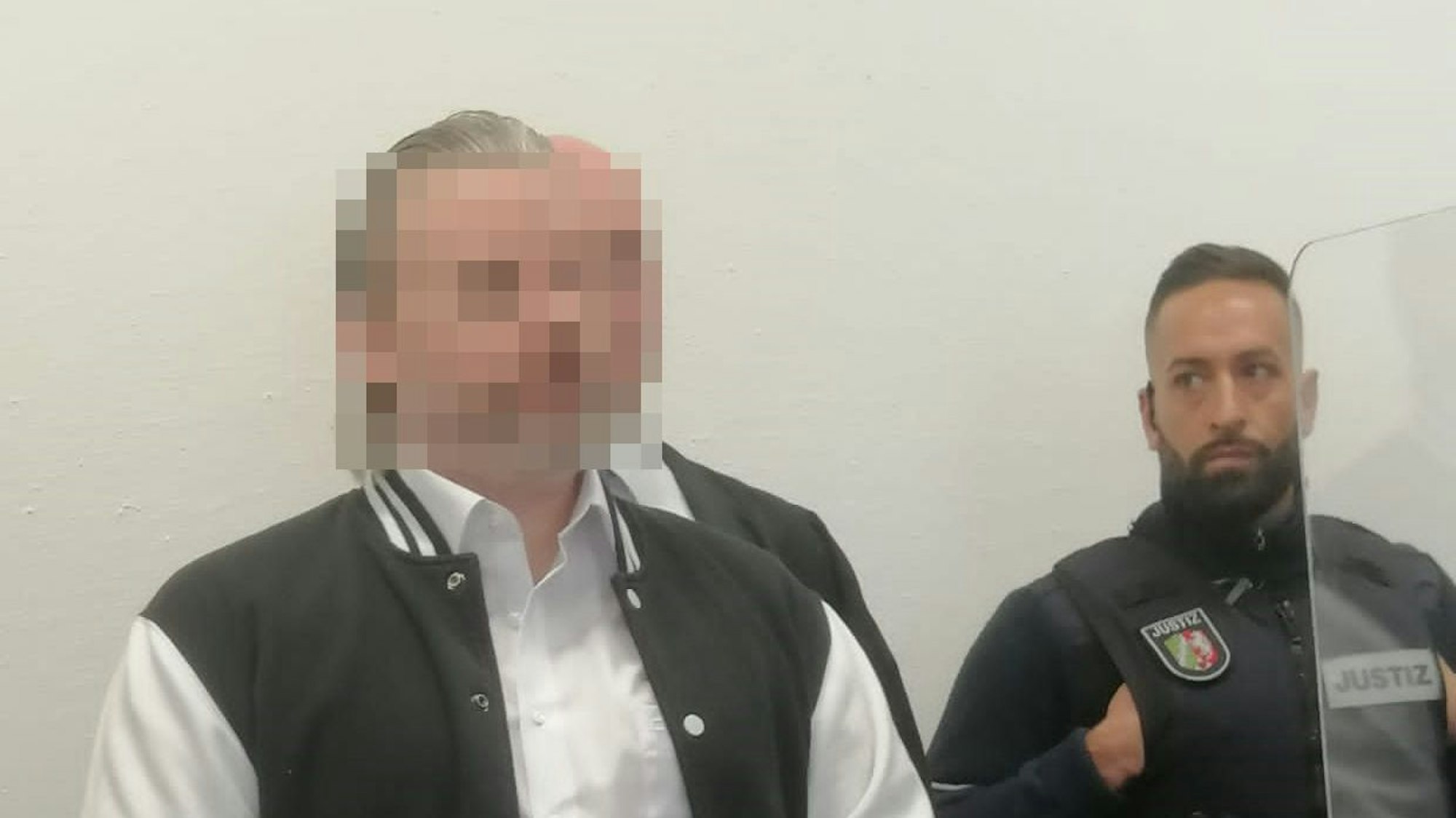 Das Bild zeigt den Angeklagten, der von einem Justizbeamten in den Gerichtssaal gebracht wird.
