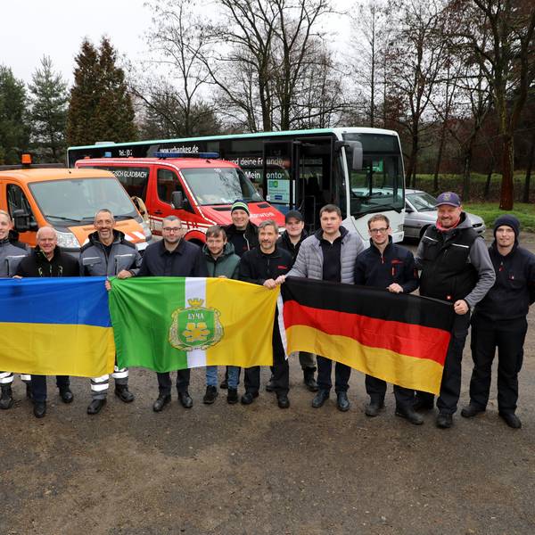 Eine Menschengruppe steht vor Bussen und hält drei Flaggen hoch.