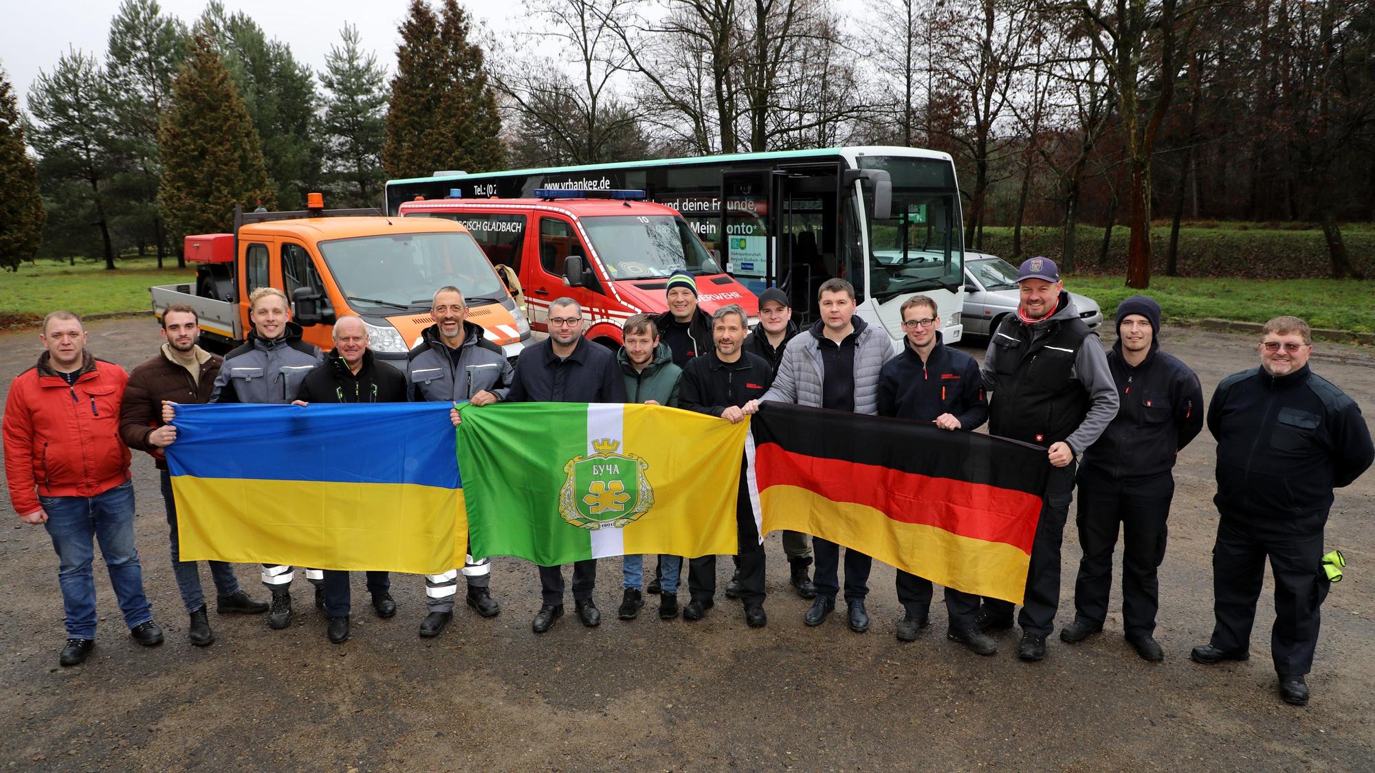 Eine Menschengruppe steht vor Bussen und hält drei Flaggen hoch.