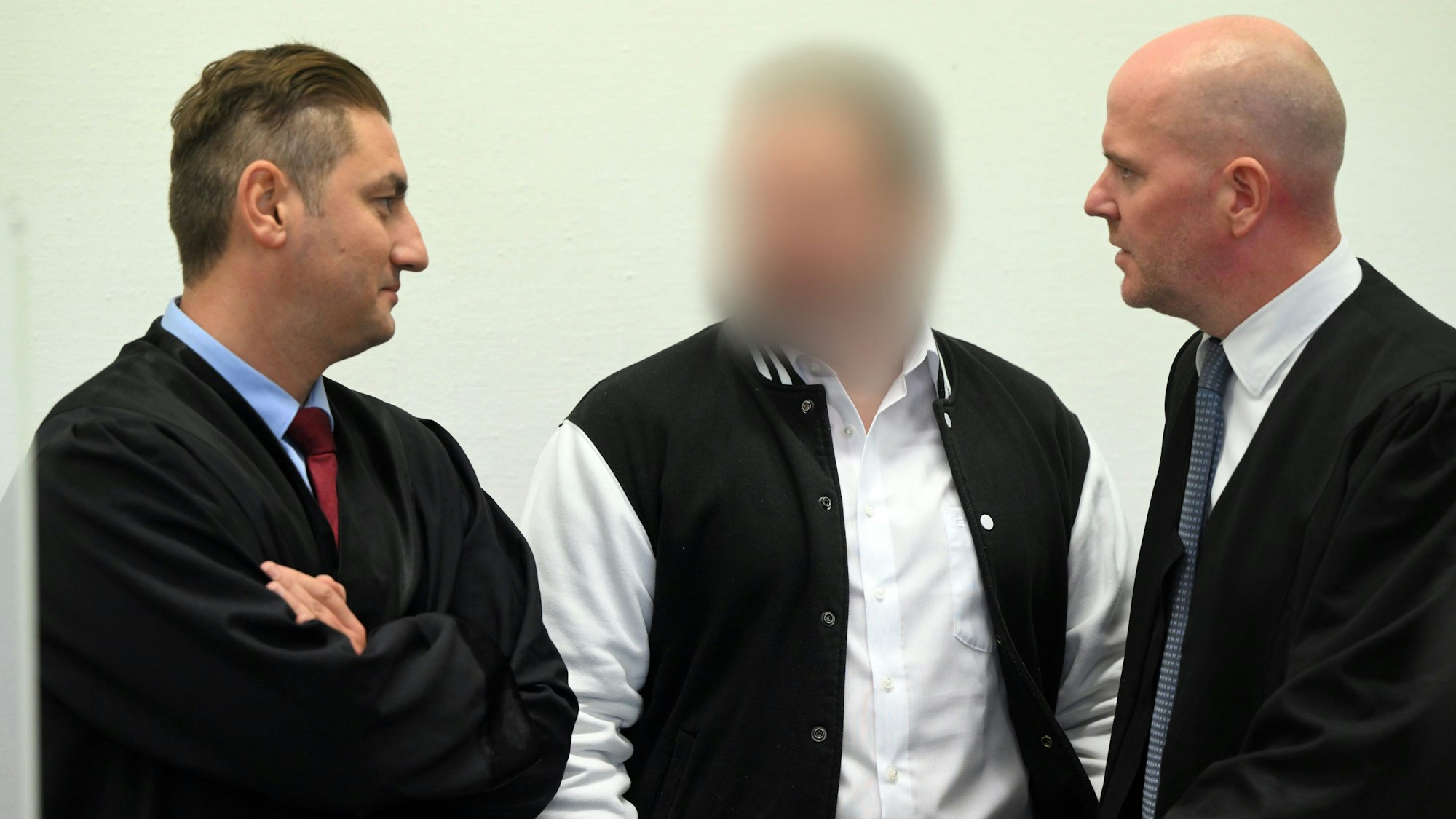 Der Angeklagte steht zwischen seinen Anwälten Mutlu Günal und Martin Bücher. Er trägt ein weißes Hemd und eine schwarz-weiße Sportjacke.