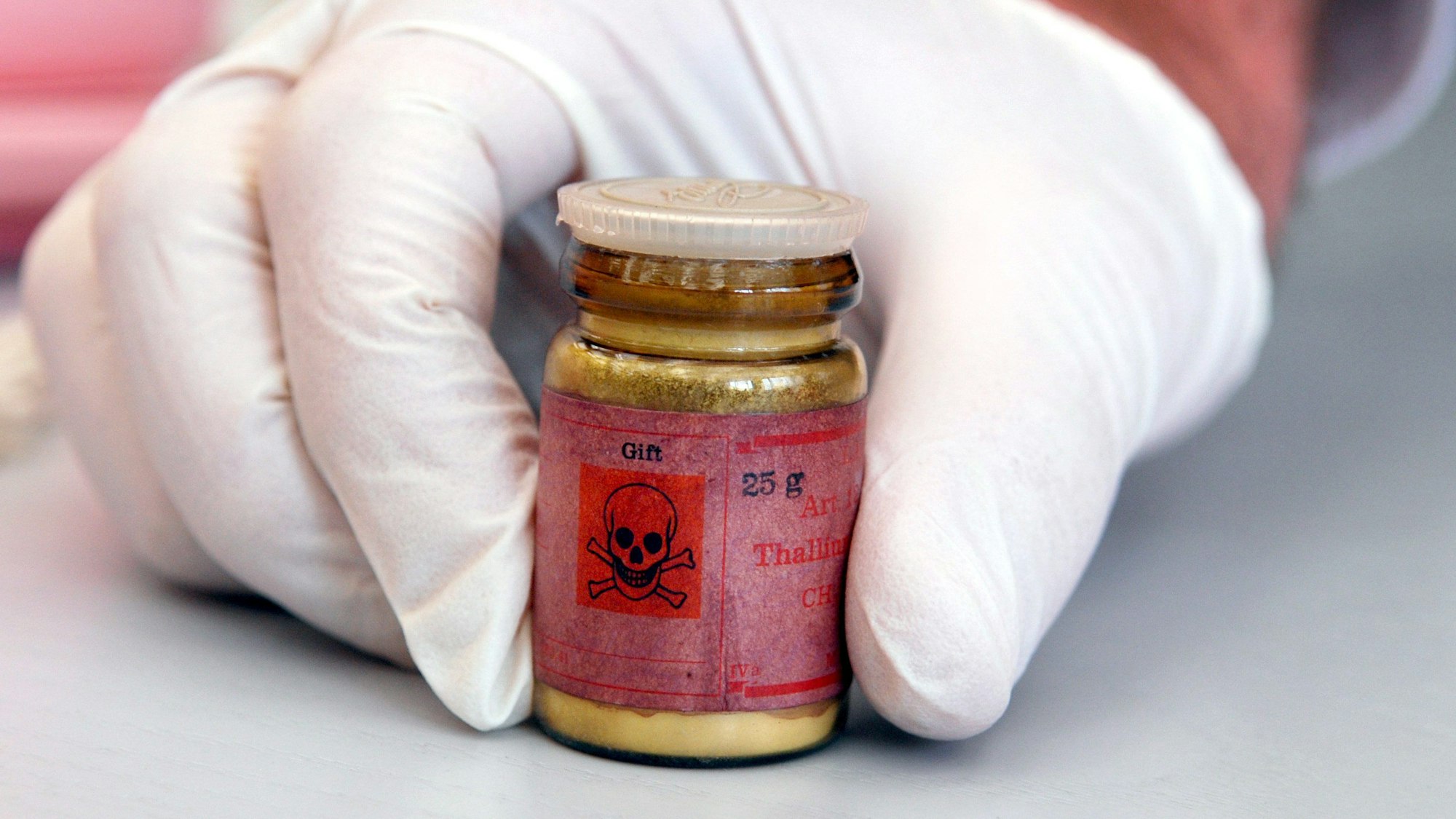 Man sieht eine behandschuhte Hand, die eine kleine Glasflasche auf einen Tisch stellt.