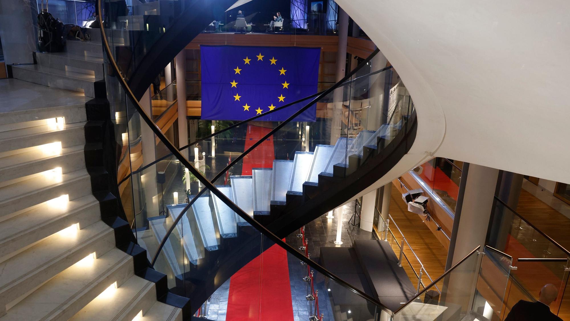 Zu sehen ist ein Treppenhaus mit zwei beleuchteten Wendeltreppen. Der Blick geht nach unten in eine Halle, in der ein roter Teppich liegt, darüber hängt die Flagge der EU.