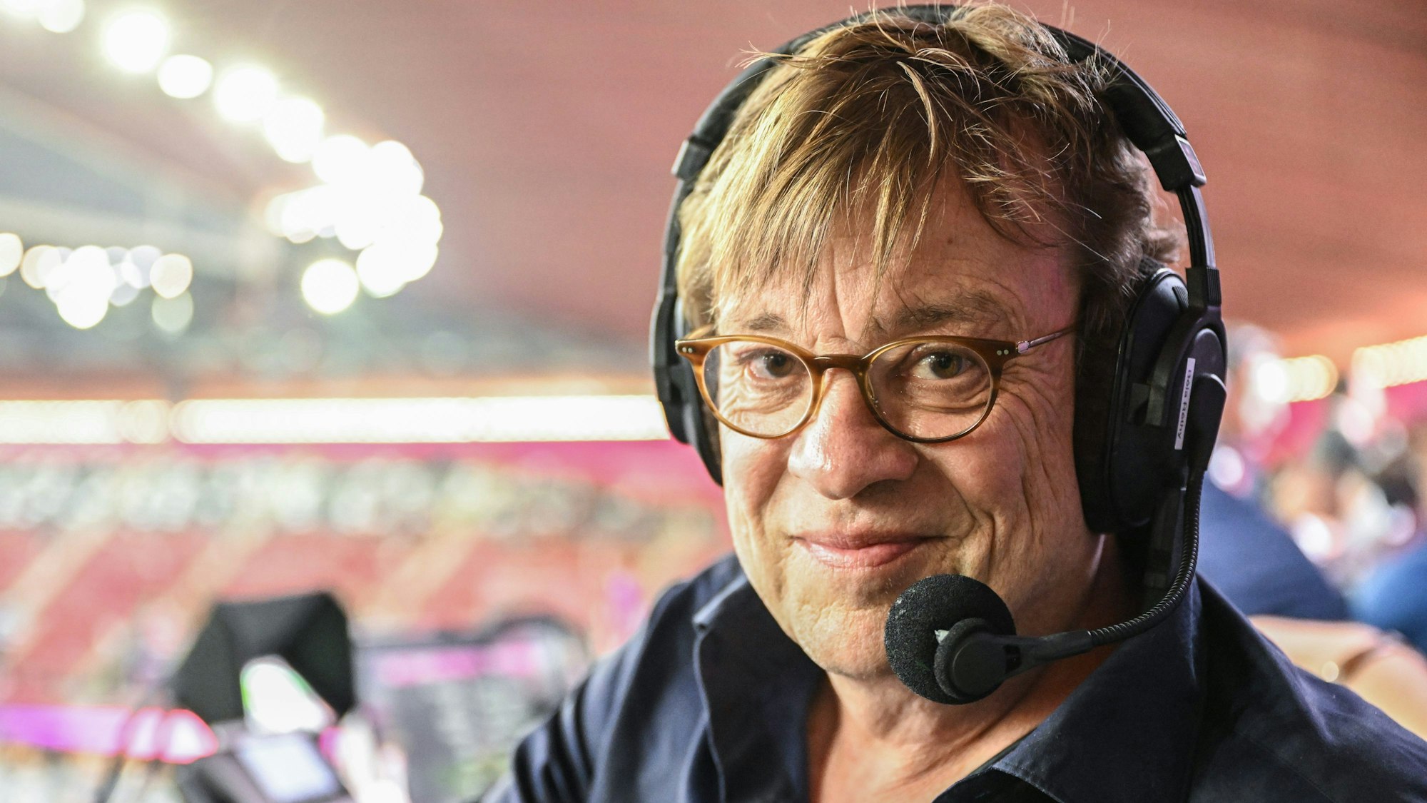 Fußball-Kommentator Béla Réthy sitzt vor einer Übertragung der Fußball-Weltmeisterschaft 2022 in Katar im Stadion an seinem Kommentatorenplatz. Er trägt ein Headset mit Mikrofon.
