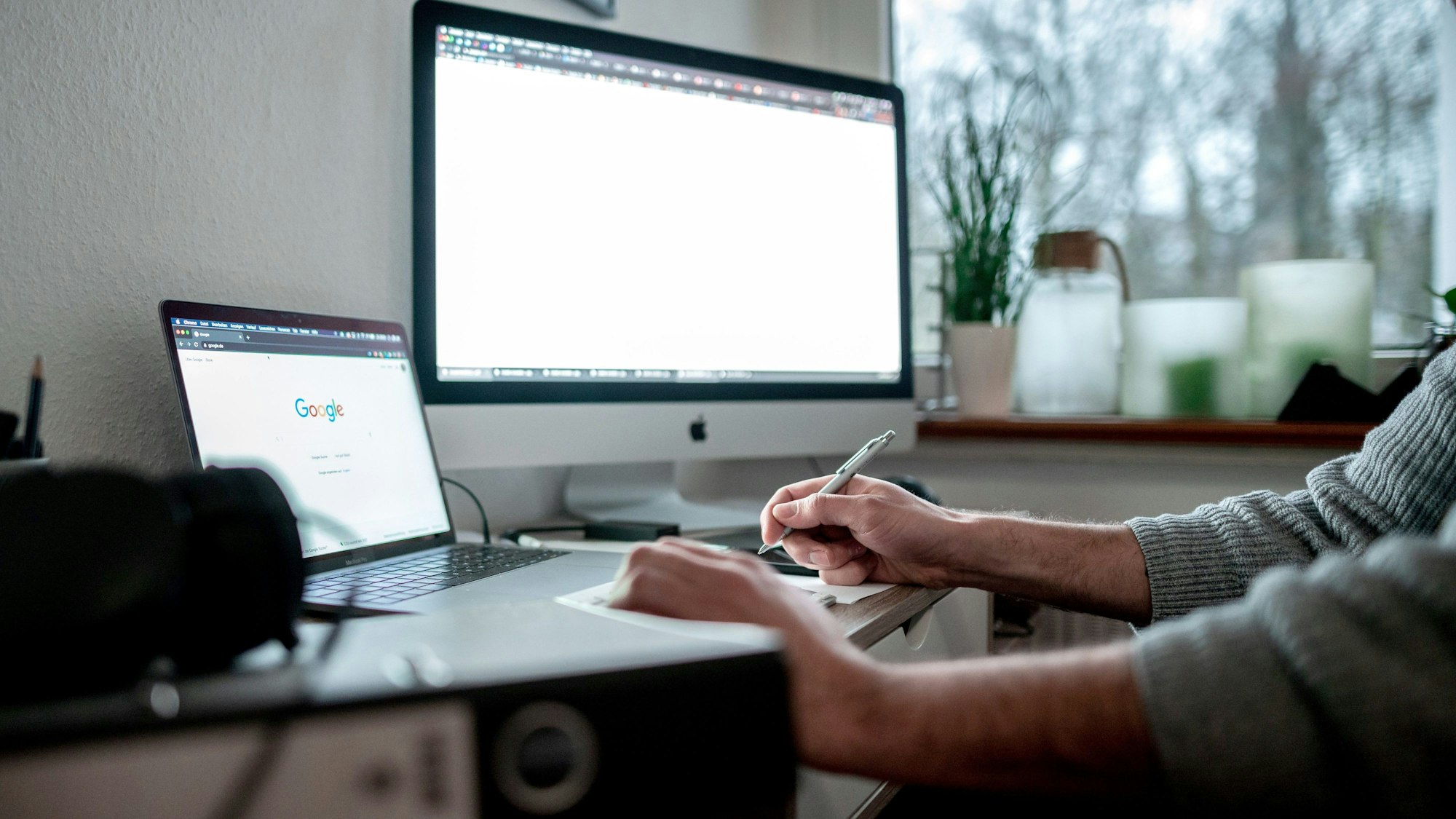 Zwei leuchtende Bildschirme stehen auf einem Schreibtisch, die Hände eines Mannes sind zu sehen.