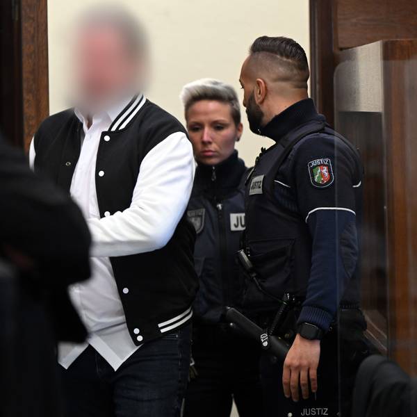 Der Angeklagte aus Hürth steht im Gerichtssaal hinter seinem Stuhl. Sein Gesicht ist verpixelt. Neben ihm steht ein Justizbeamter.