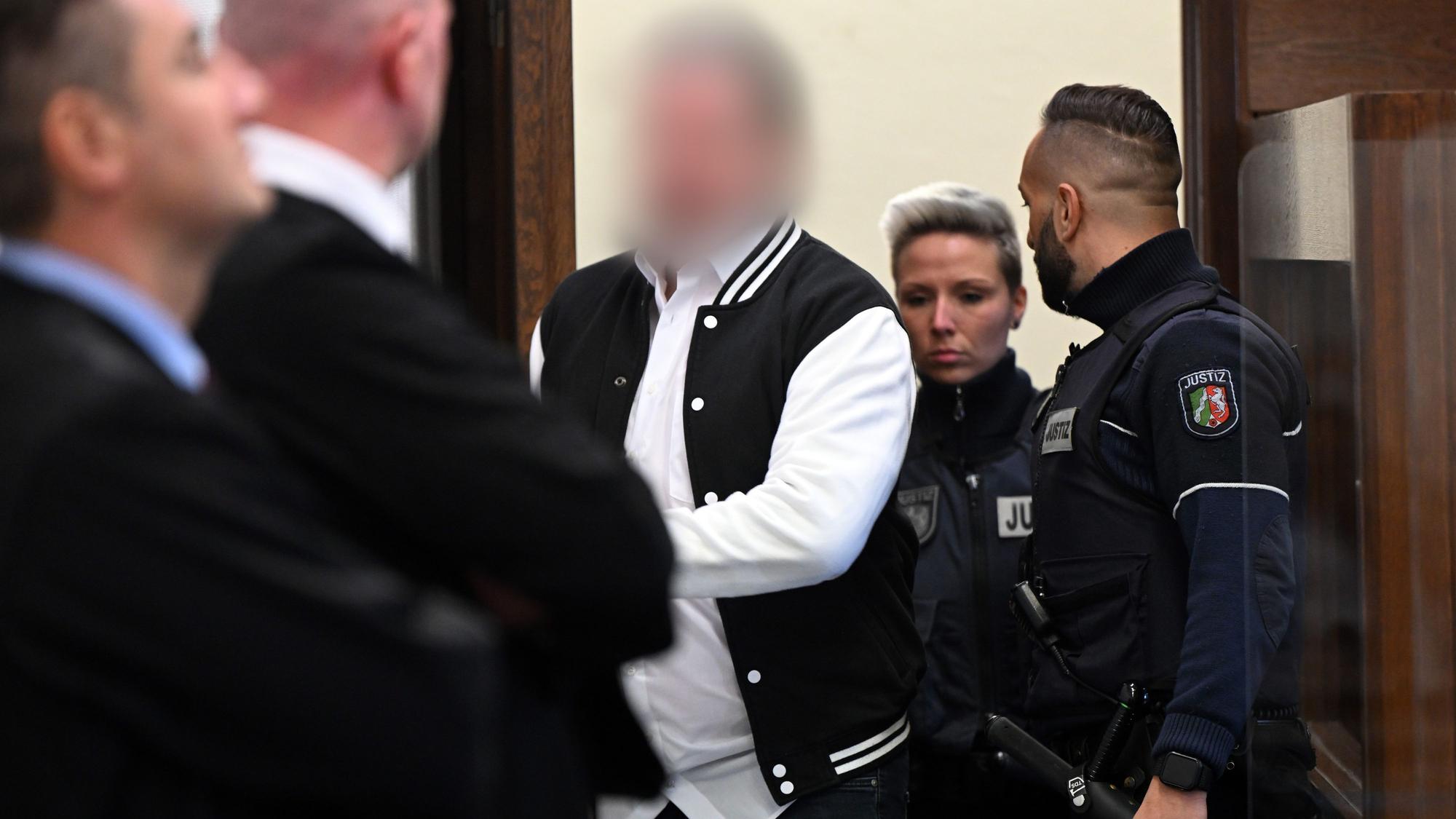Der Angeklagte aus Hürth steht im Gerichtssaal hinter seinem Stuhl. Sein Gesicht ist verpixelt. Neben ihm steht ein Justizbeamter.