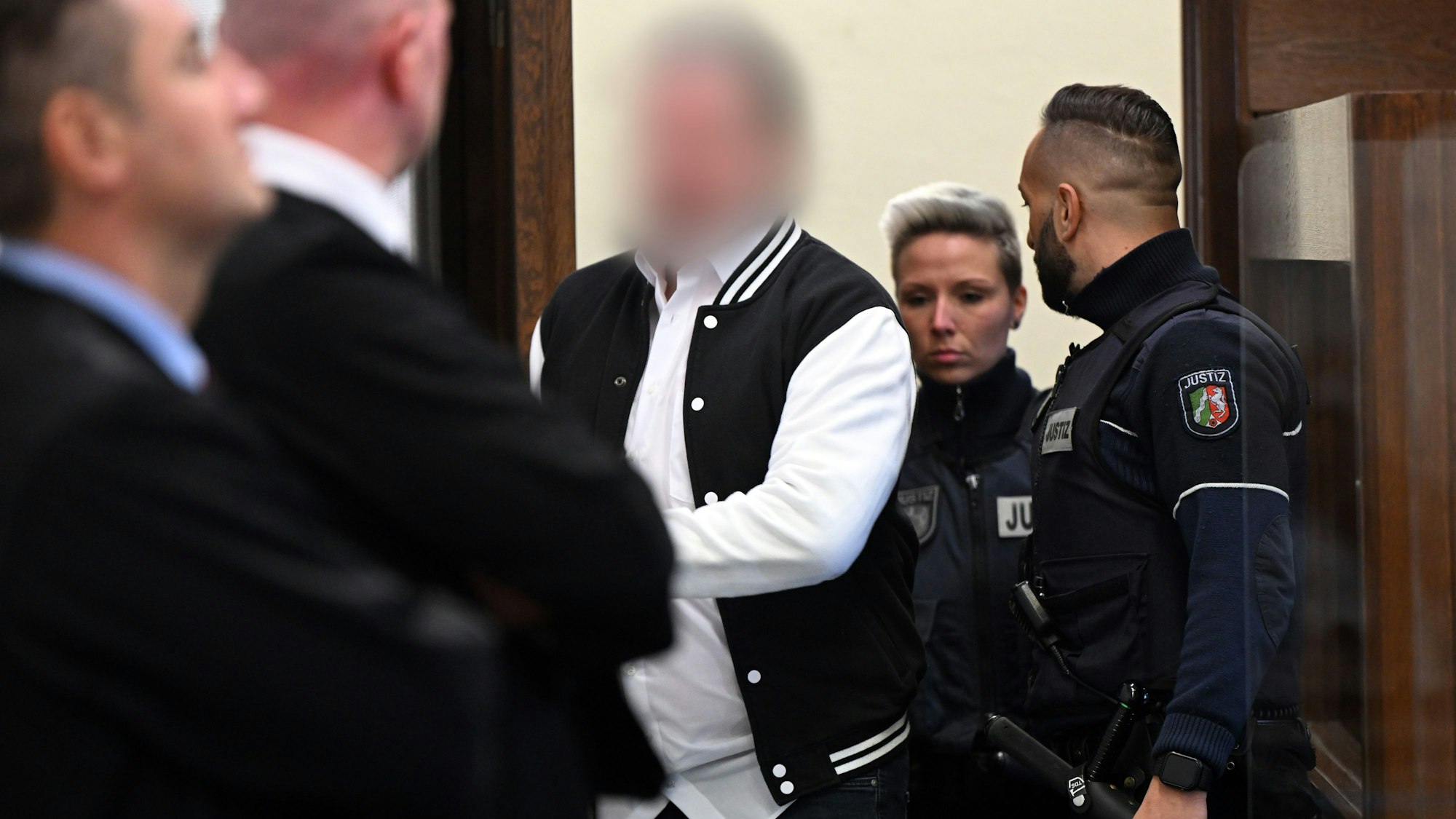 Der Angeklagte aus Hürth steht im Gerichtssaal hinter seinem Stuhl. Sein Gesicht ist verpixelt. Neben ihm steht ein Justizbeamter.