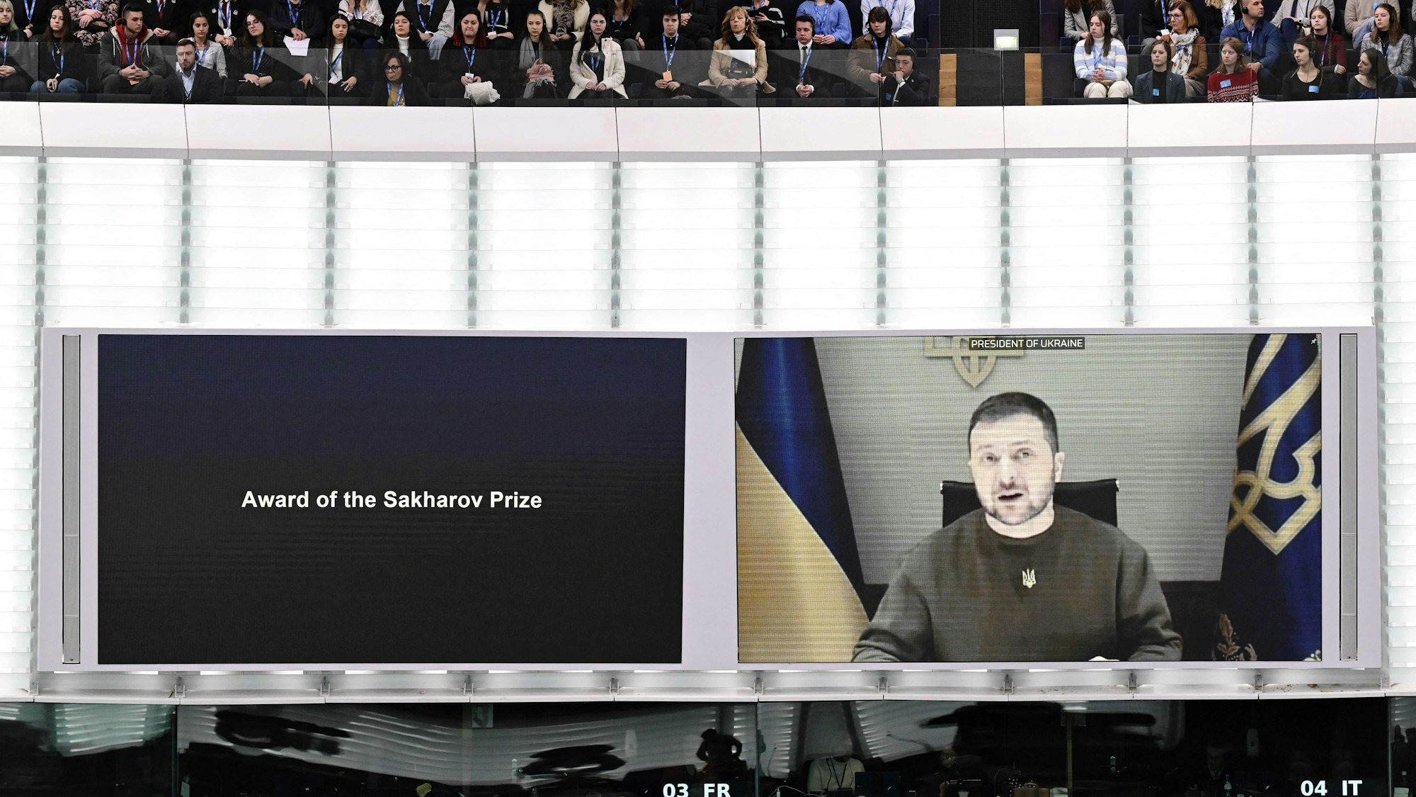 Man sieht zwei Bildschirme an der Wand, auf dem rechten ist Präsident Selenskyj, auf dem linken steht „Award of the Sakharov Price“. Über den Bildschirmen sieht man Personen auf einer Tribüne.