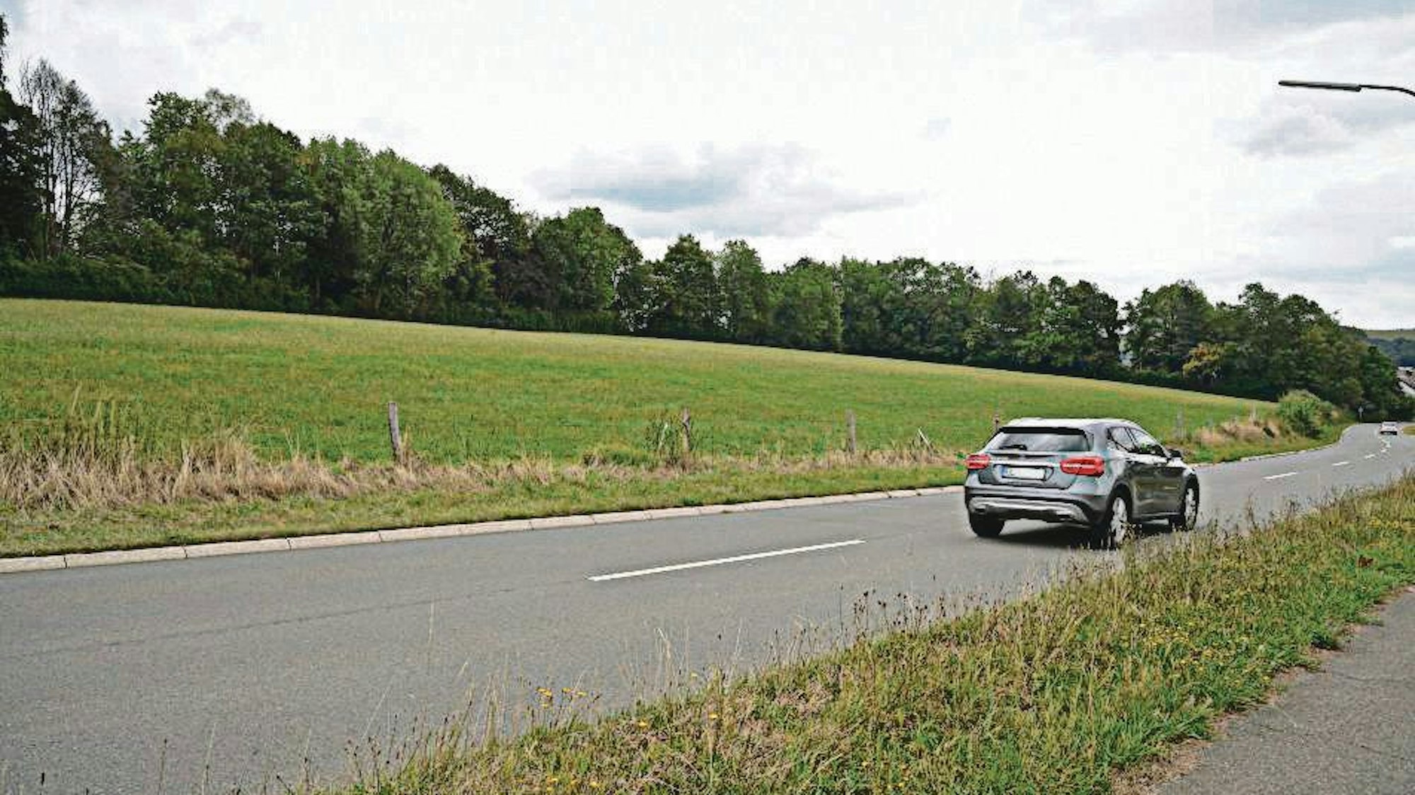 Ein silbernes Auto fährt auf einer Straße, links eine Wiesenfläche, wo das neue Baugebiet in Kürten-Busch entstehen soll.