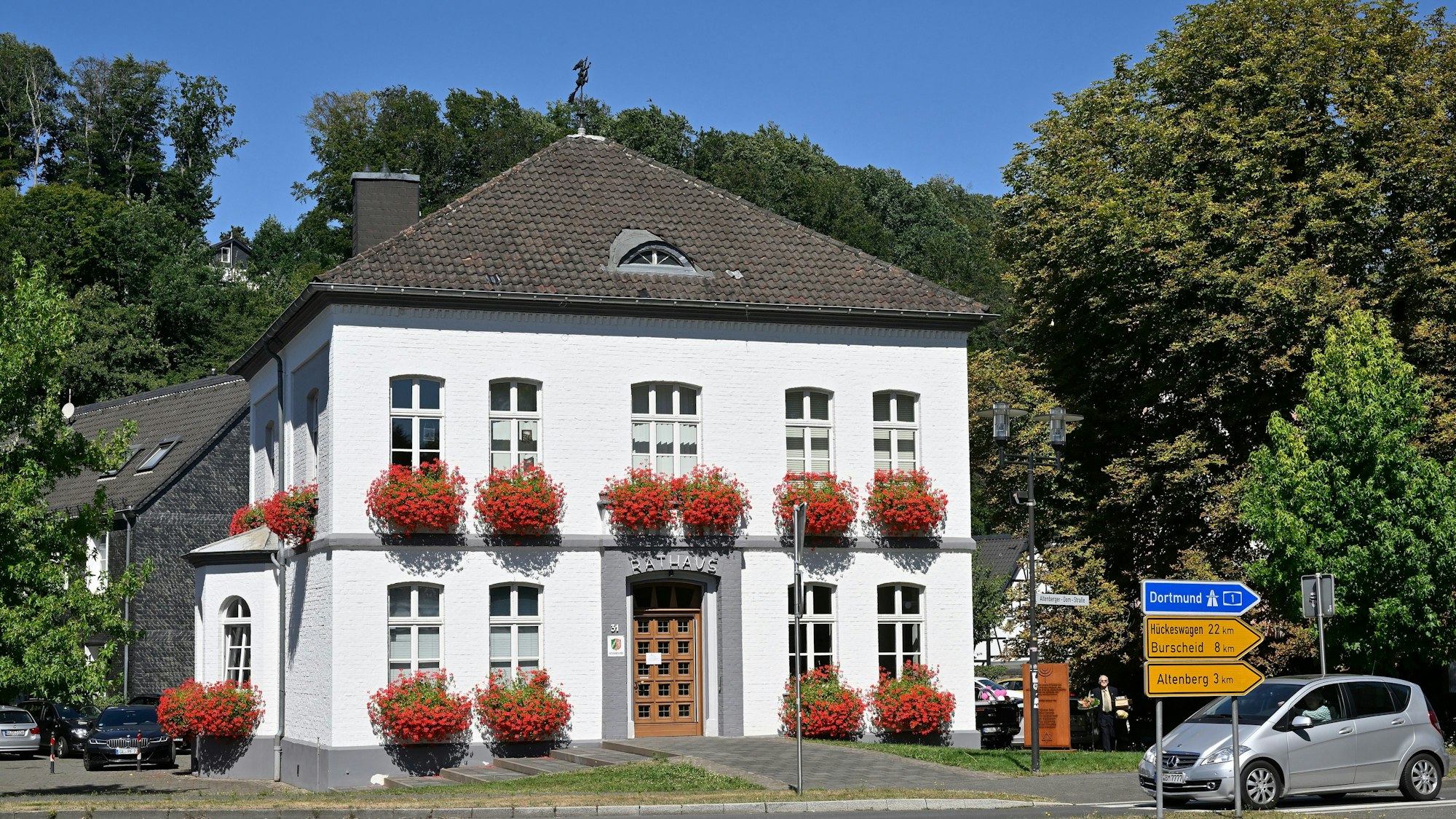 Am Odenthaler Rathaus hängen Blumenkästen. Im Hintergrund stehen Bäume.