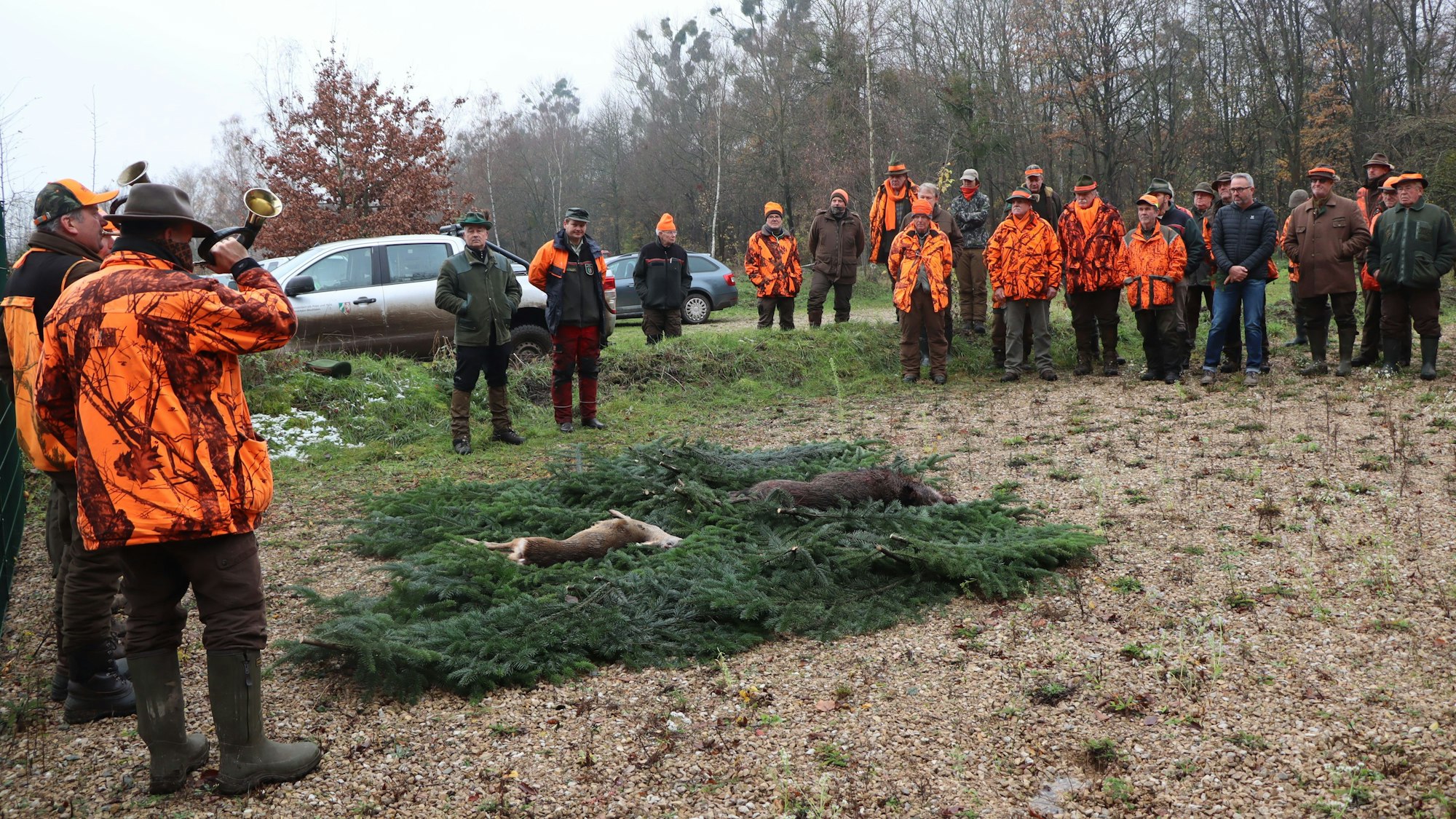 Auf Tannenzweigen liegen ein geschossenes Wildschwein und ein Reh. Im Halbkreis haben sich die Jäger um die toten Tiere versammelt.