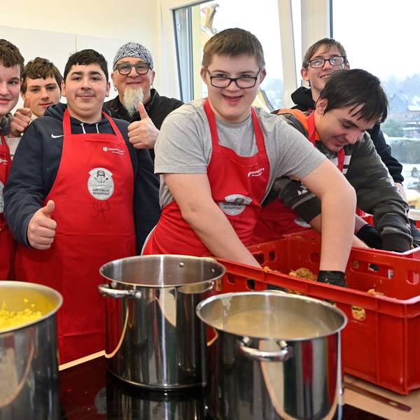 Schüler tragen rote Kochschürzen und stehen in der Schulküche um Sternekoch Stefan Marquard herum.