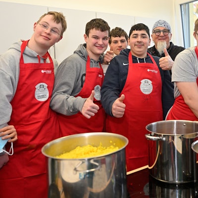 Schüler tragen rote Kochschürzen und stehen in der Schulküche um Sternekoch Stefan Marquard herum.