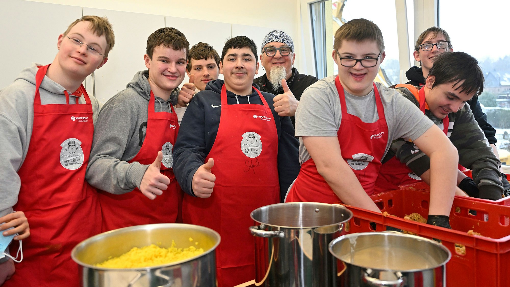 Schüler tragen rote Kochschürzen und stehen in der Schulküche um Sternekoch Stefan Marquard herum.