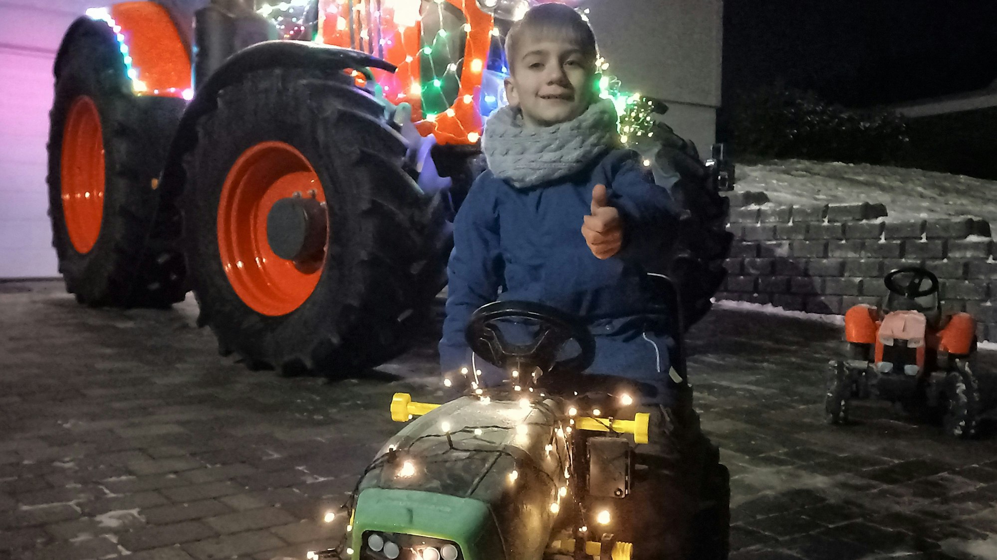Blick auf einen Jungen auf einem mit Lichtern geschmückten Spieltraktor. Im Hintergrund ein mit Lichtern geschmückter großer Traktor.