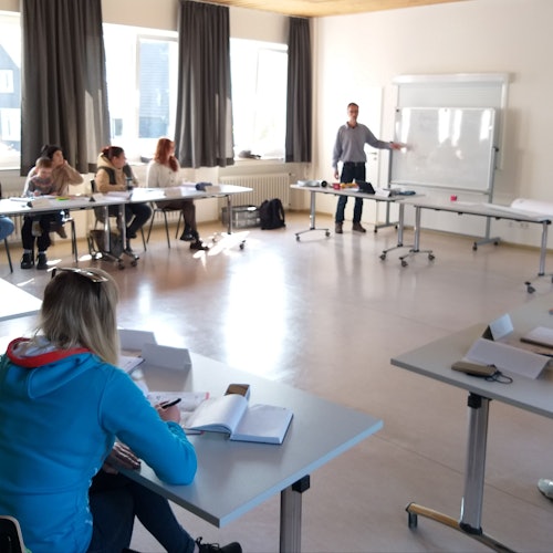 Frauen sitzen in einem Raum an Tischen und hören dem Sprachlehrer zu, der vorn an einer weißen Tafel etwas erklärt.