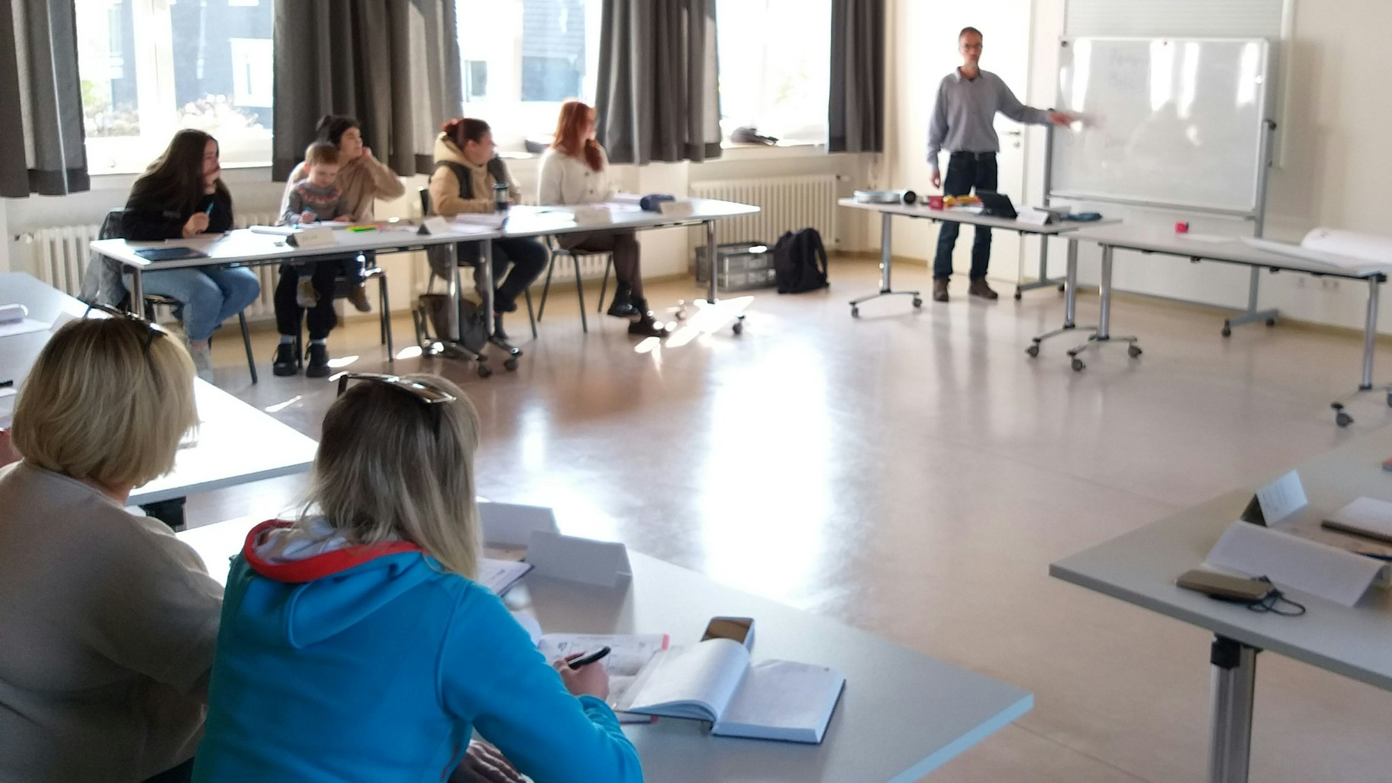 Frauen sitzen in einem Raum an Tischen und hören dem Sprachlehrer zu, der vorn an einer weißen Tafel etwas erklärt.