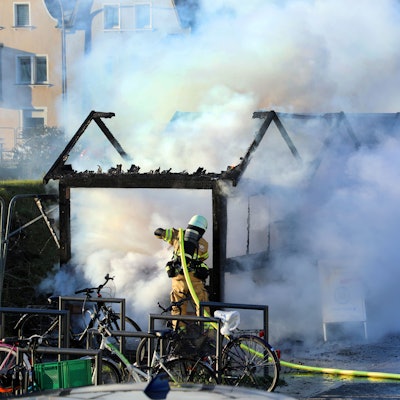Zwei Feuerwehrleute löschen ein brennendes Holzhäuschen. Große Rauchwolken steigen auf.