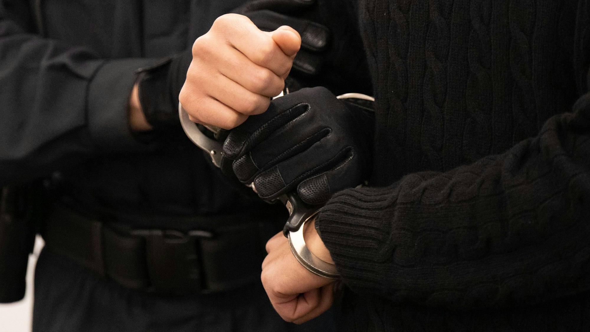 Man sieht, wie einer Person von einer anderen Person Handschellen angelegt werden. Beide Personen tragen schwarze Kleidung, die Person ohne Handschellen trägt schwarze Handschuhe.