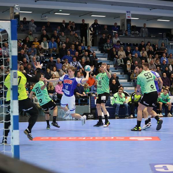Man sieht eine Spielsituation, der junge Handballer Finn Schroven setzt zum Wurf auf das Tor an. Um ihn herum stehen Spieler in anderen Trikots, im Hintergrund sieht man die Tribüne.