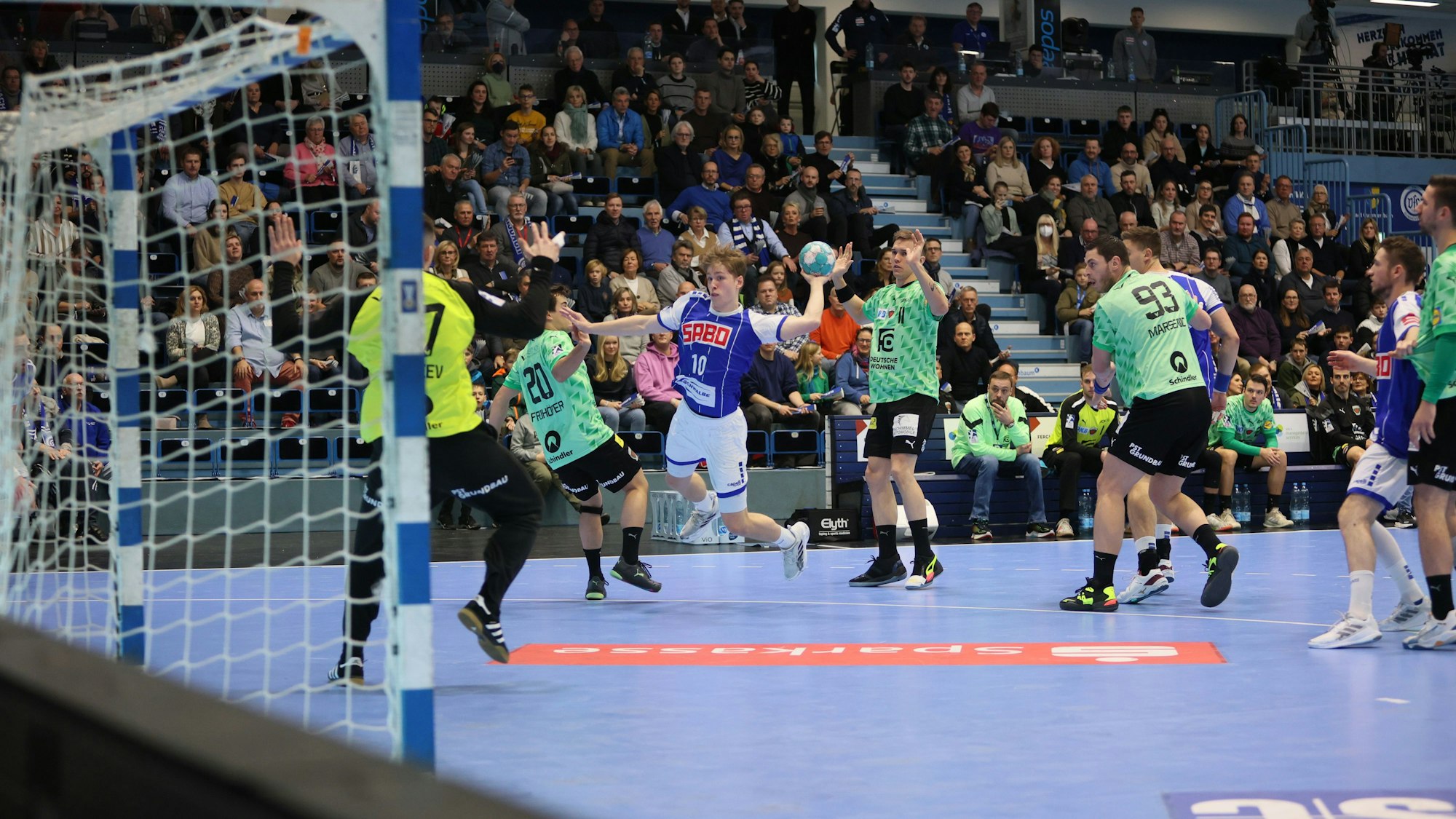 Man sieht eine Spielsituation, der junge Handballer Finn Schroven setzt zum Wurf auf das Tor an. Um ihn herum stehen Spieler in anderen Trikots, im Hintergrund sieht man die Tribüne.