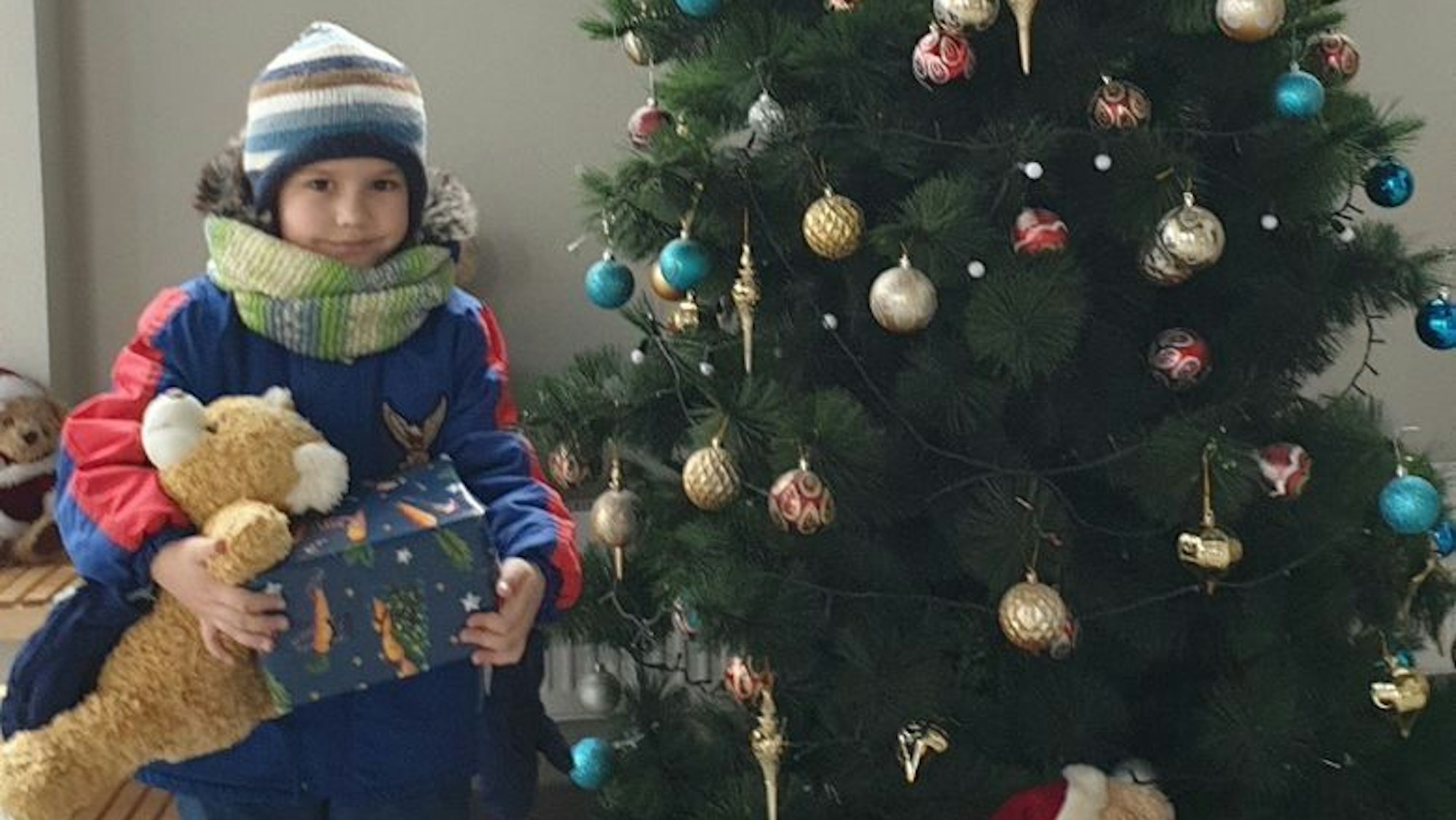 Ein Kind steht mit Kuscheltier und Geschenk neben einem geschmückten Baum.