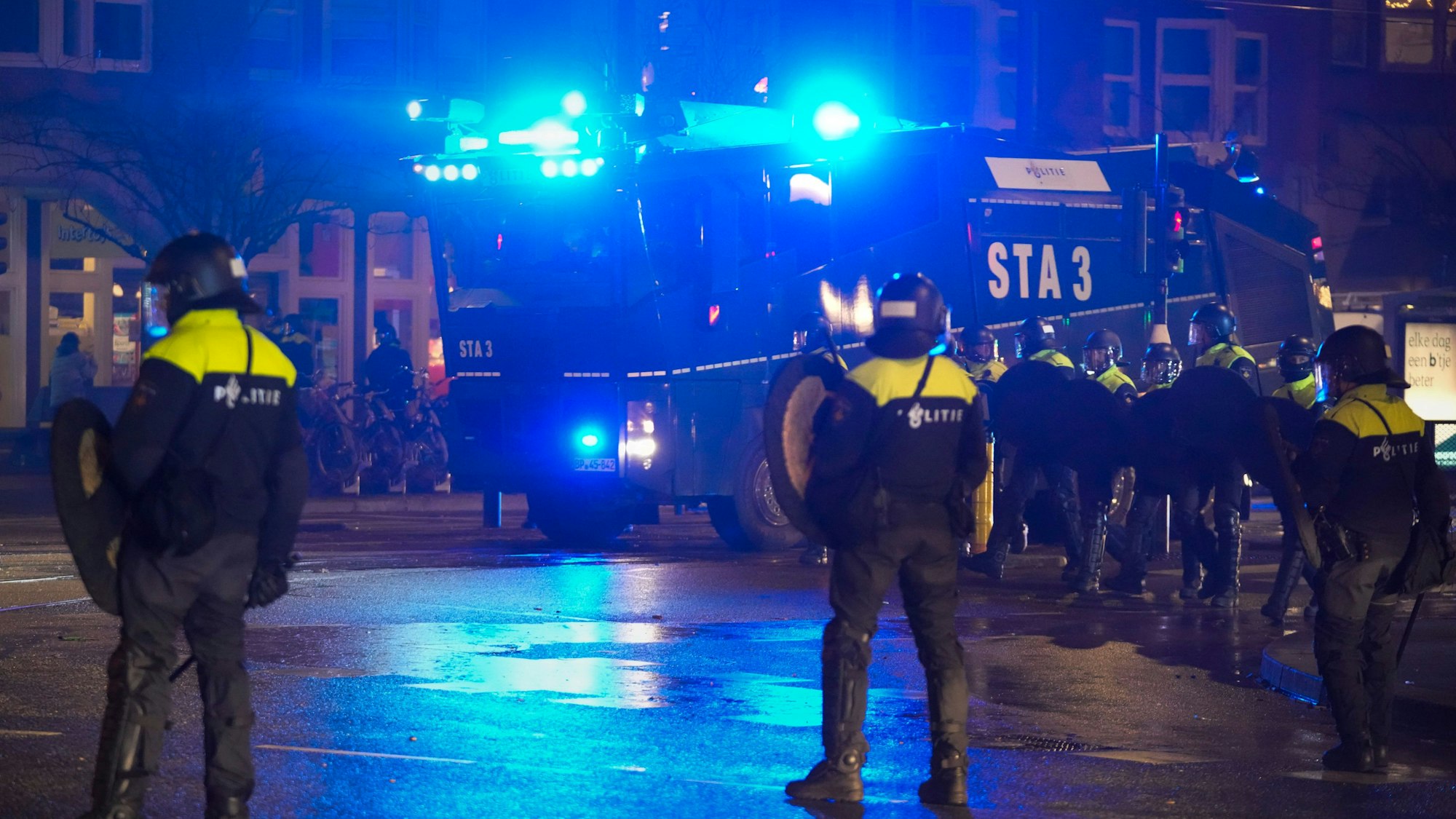 Die niederländische Polizei steht bei Dunkelheit auf einer großen Straße in Amsterdam, im Hintergrund steht ein Wasserwerfer der Bundespolizei aus Sankt-Augustin.