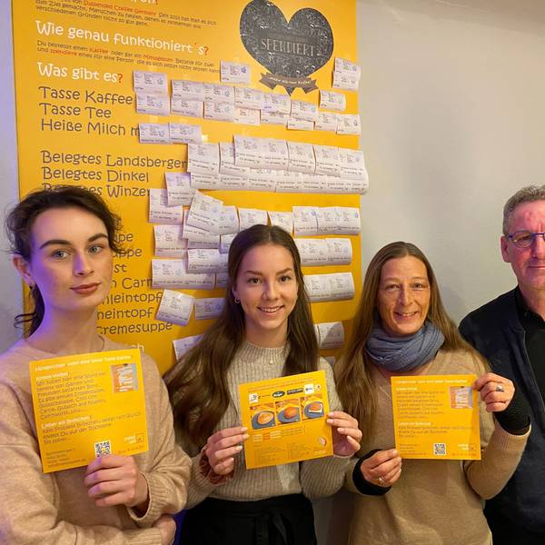 Anna Kutschenko, Lilly Rausch, Bianca Dressler und Antonius Quodt stehen im Café „Status Quodt“ vor der gelben Tafel, an der bezahlte Bons mit Getränken und Speisen angebracht sind.