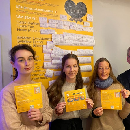 Anna Kutschenko, Lilly Rausch, Bianca Dressler und Antonius Quodt stehen im Café „Status Quodt“ vor der gelben Tafel, an der bezahlte Bons mit Getränken und Speisen angebracht sind.