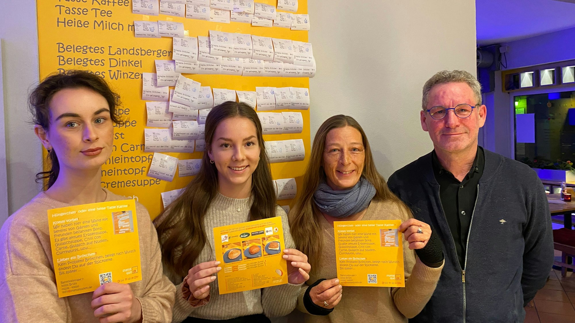 Anna Kutschenko, Lilly Rausch, Bianca Dressler und Antonius Quodt stehen im Café „Status Quodt“ vor der gelben Tafel, an der bezahlte Bons mit Getränken und Speisen angebracht sind.