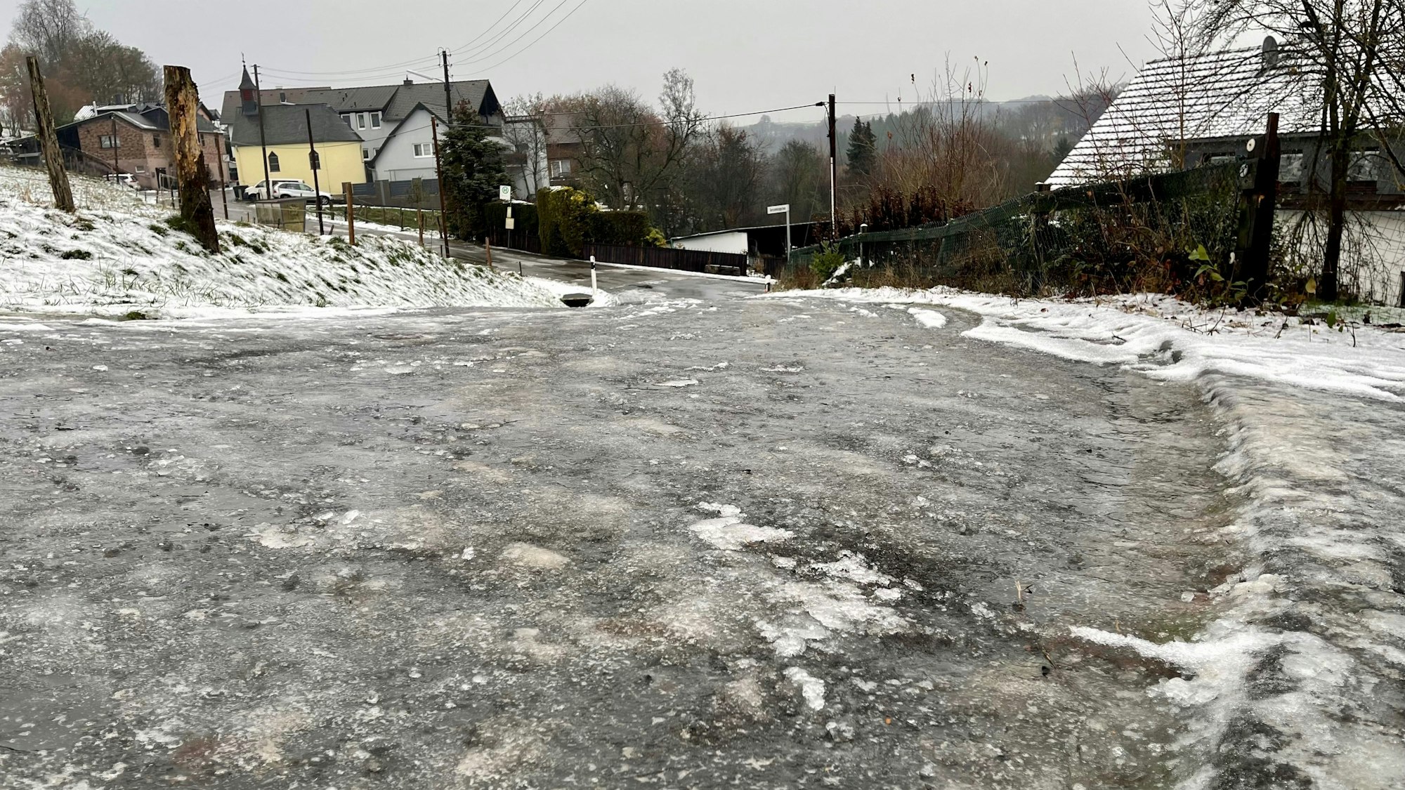 Eine zentimeterdicke Eisschicht liegt auf einer abschüssigen Straße im Hennefer Ortsteil Hanf. Im Hintergrund sind Häuser zu sehen.