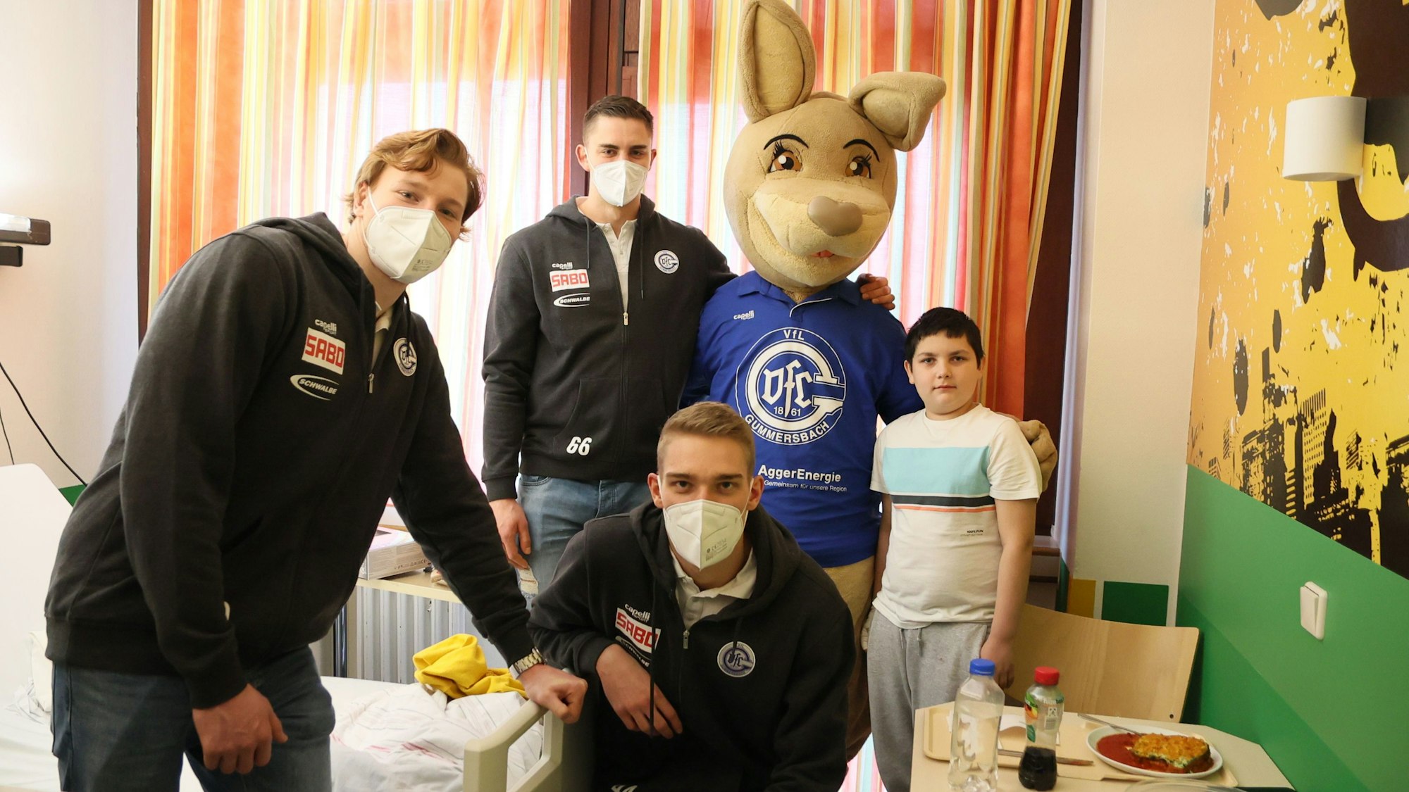 Die Handballspieler Tom Kiesler, Stepan Zeman und Tom Jansen posieren mit dem lebensgroßen VfL-Maskottchen, dem Känguru Gummi und dem zehn Jahre alten Ben für ein Gruppenfoto in einem Krankenzimmer der Kinderklinik.