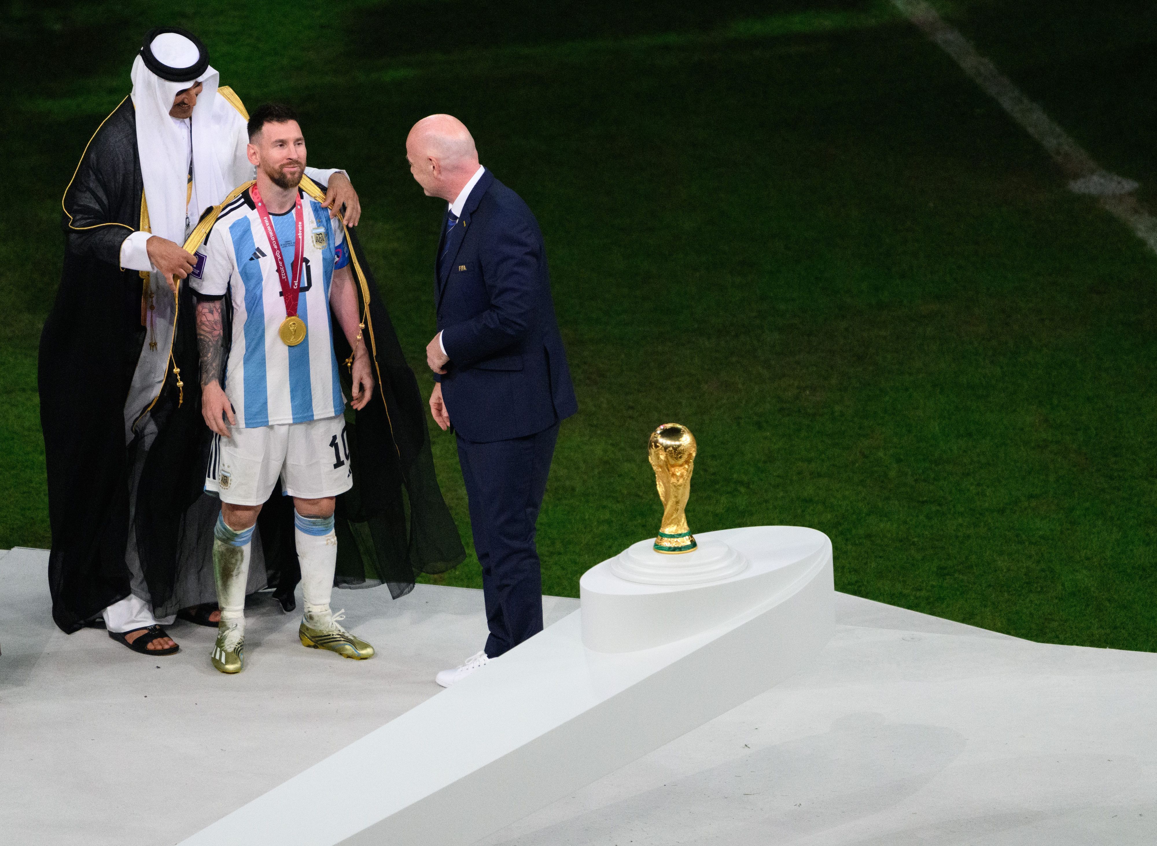 Der Emir von Katar, Scheich Tamim bin Hamad Al Thani (l), legt Argentiniens Lionel Messi vor Übergabe des WM-Pokals vor Fifa-Präsident Gianni Infantino ein Bischt, ein traditionelles arabische Kleidungsstück an.