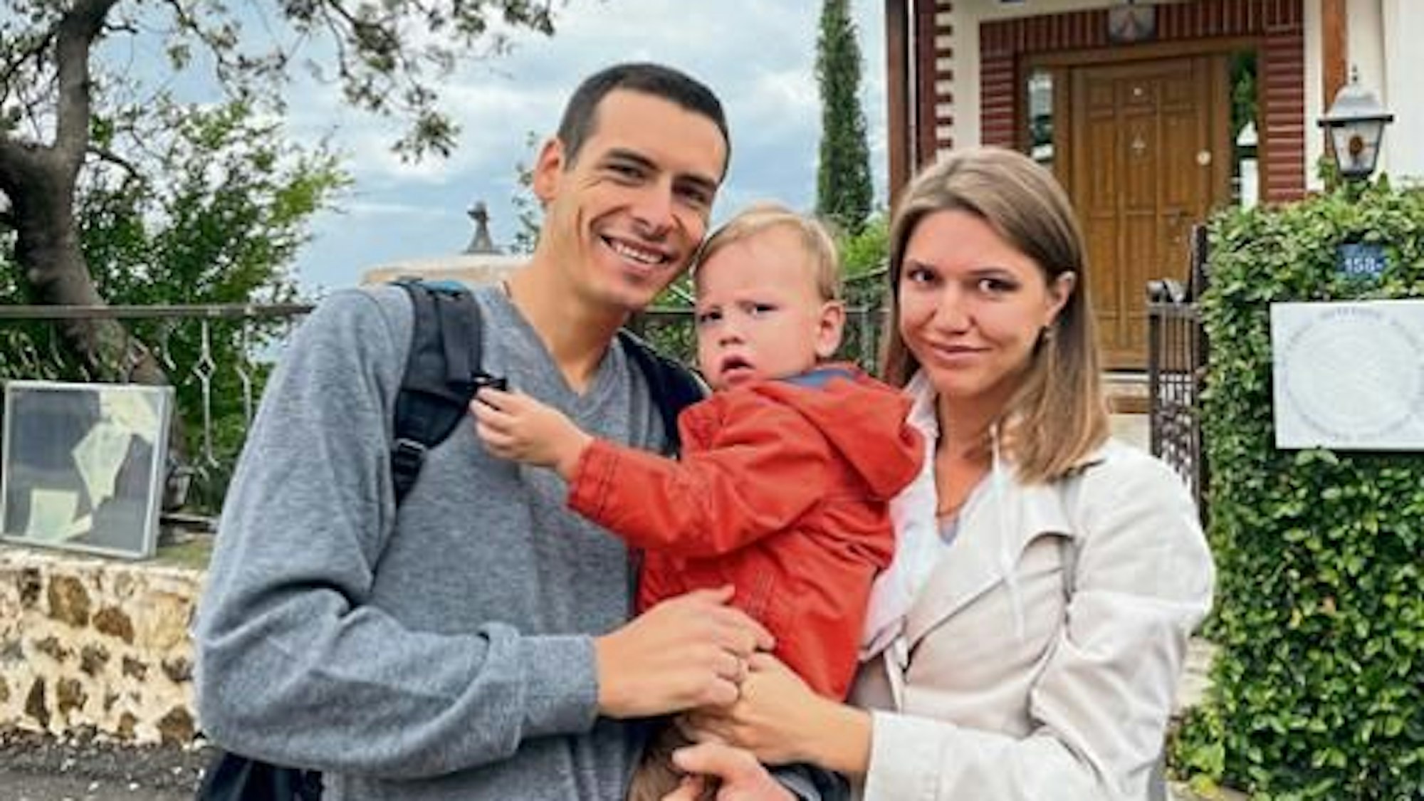 Ein russisch-ukrainisches Paar steht mit ihrem Sohn im Arm auf der Straße und lächelt in die Kamera.