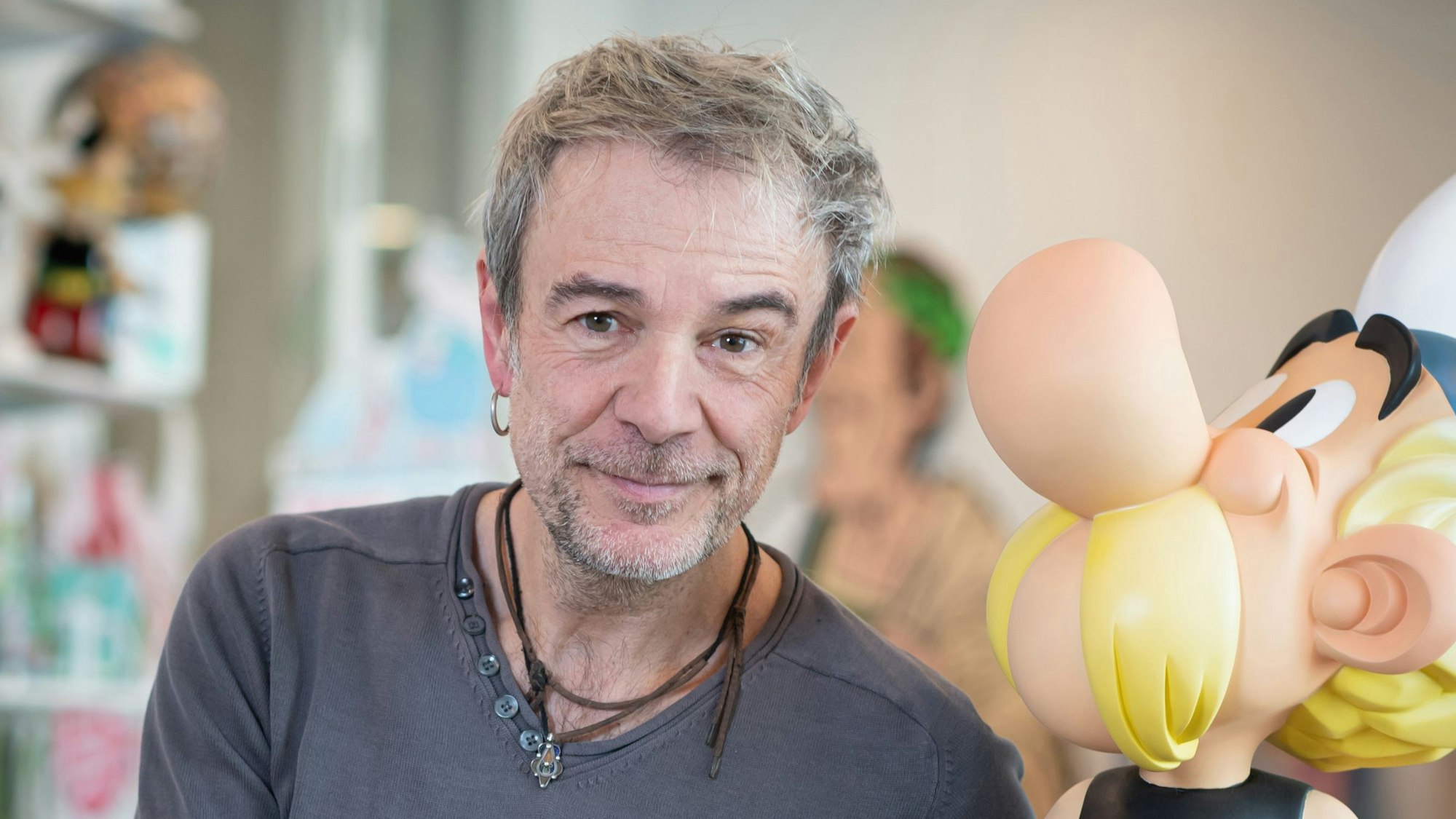 Texter Fabrice Caro sitzt neben der Figur Asterix und lächelt in die Kamera.