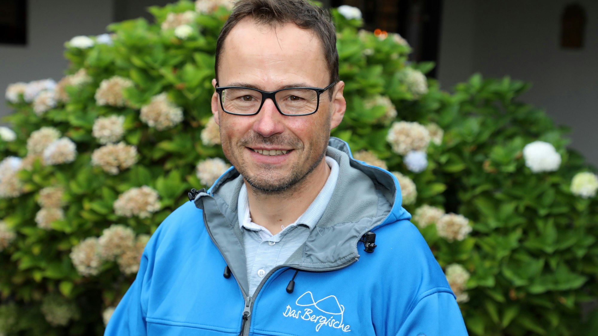 Tobias Kelter steht mit einer Jacke von „Das Bergische“ vor einem Strauch.