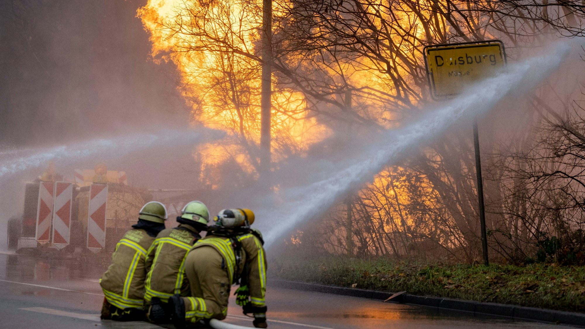 Feuerwehrleute arbeiten daran, das brennende Gasleck in Duisburg zu löschen. Ein Feuer und Rauchschwaden sind zu sehen, im Vordergrund die Feuerwehrleute.