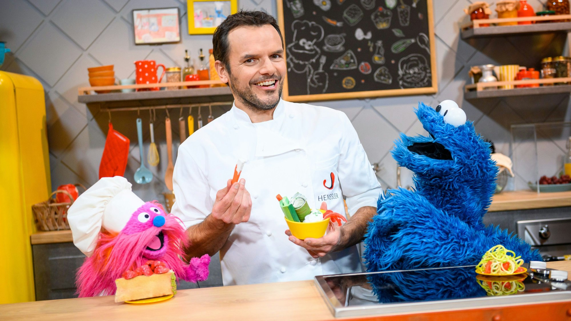 Fernsehkoch Steffen Henssler zwischen den Puppen „Gonger“ (l) und „Krümelmonster“ am Film-Set. Er hält Zutaten in der Hand und lächelt. Die Puppen schauen ihn begeistert an