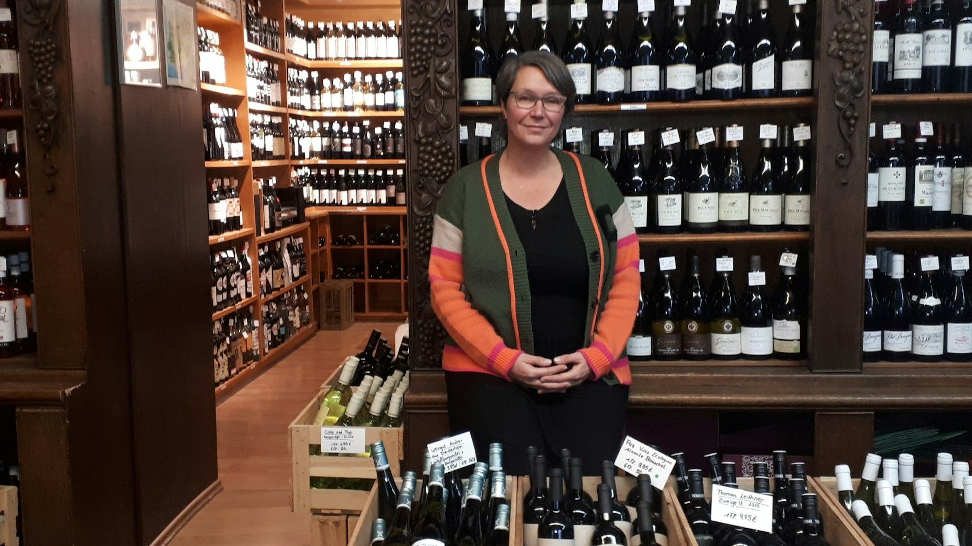Eine 50 Jahre alte Frau mit dunklen Haaren steht in einem Weinladen. Hinter und vor ihr sind Regale und Kisten mit Weinflaschen.