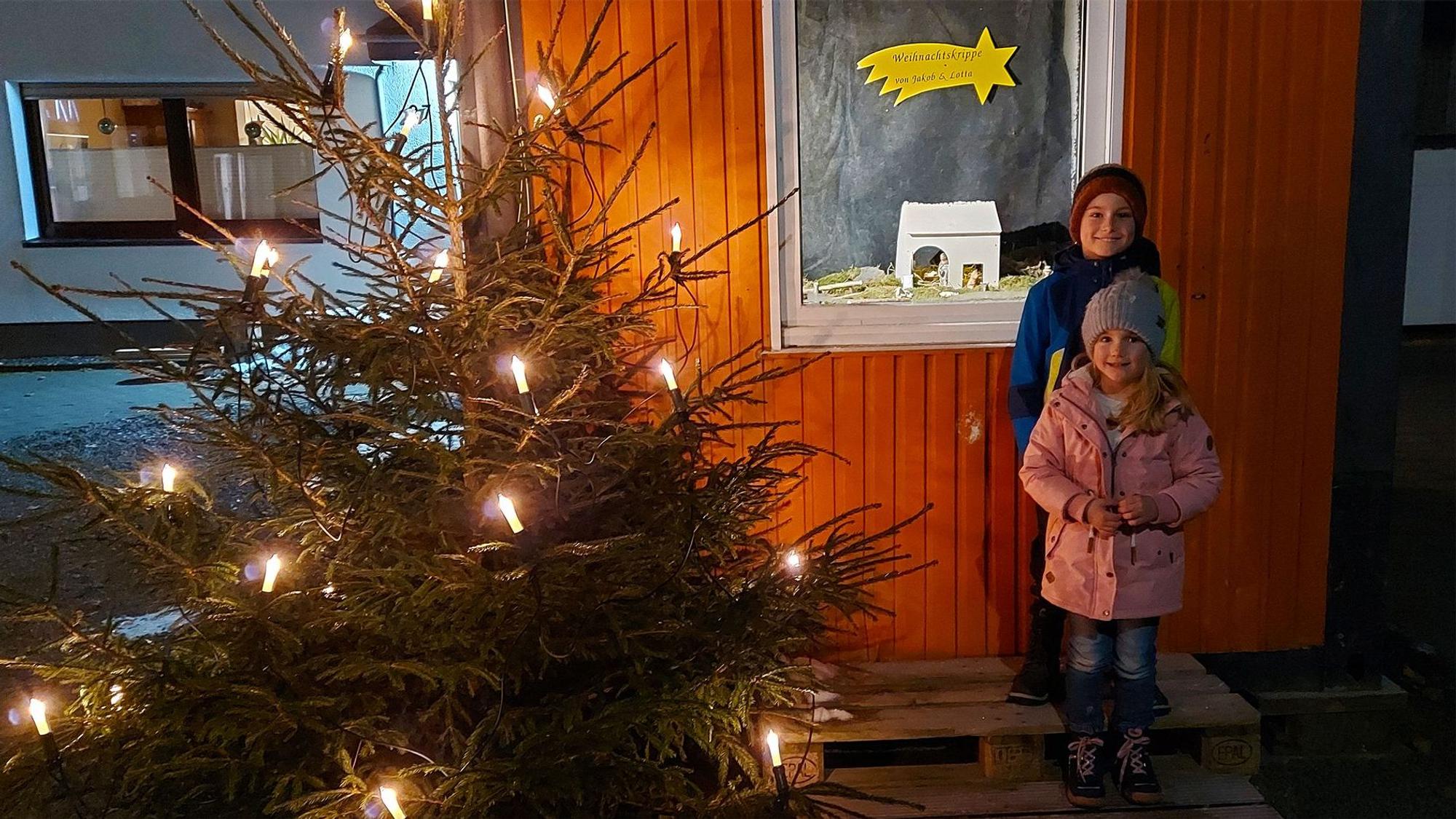 Zwei Kinder stehen neben einem Weihnachtsbaum an einem Fenster mit einer Krippe.