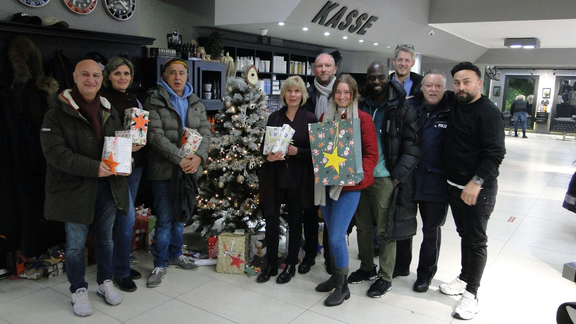 Zehn Personen stehen teils mit hübsch verpackten Geschenken in der Hand um einen Weihnachtsbaum herum, der in einem Friseur-Salon steht.