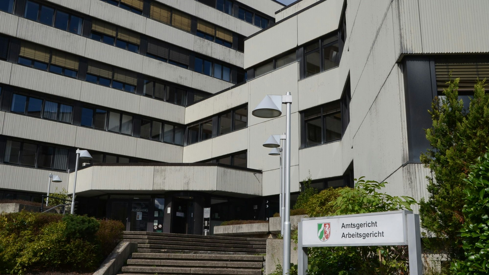Eine Treppe, von Laternen eingerahmt, führt zum Eingang eines weißen Gebäudes mit vielen Fenstern. Auf einem Schild mit dem Wappen von Nordrhein-Westfalen stehen die Bezeichnungen Amtsgericht und Arbeitsgericht.