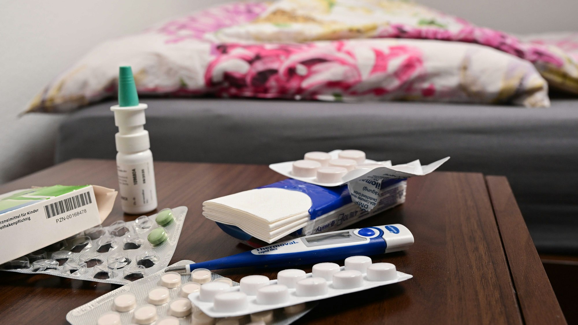 Ein Fieberthermometer und Medikamente liegen auf einem Nachttisch, im Hintergrund ein Bett. (Symbolbild)