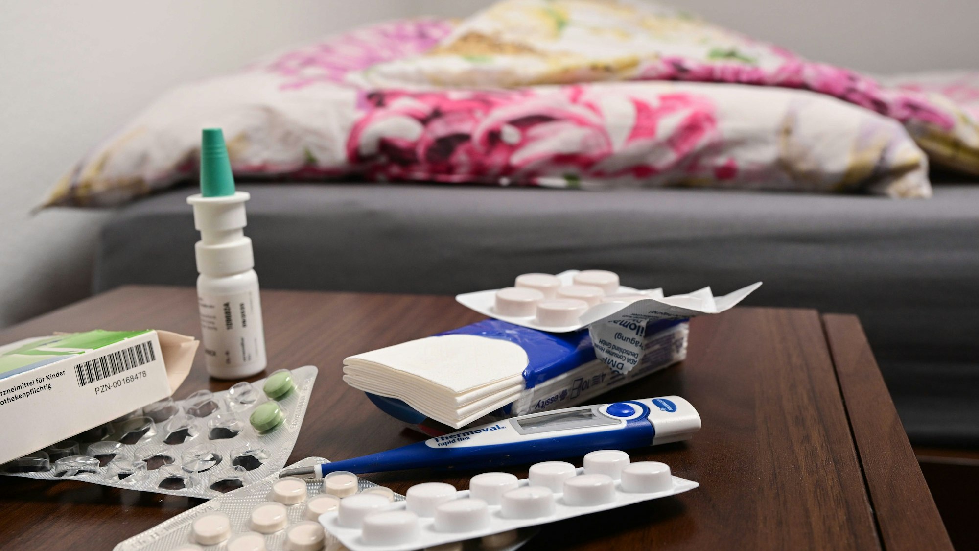 Ein Fieberthermometer und Medikamente liegen auf einem Nachttisch.
