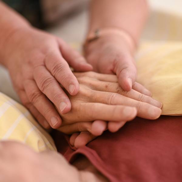 Eine Person hält mit beiden Händen die Hand eines Menschen, der mit einer gelben Decke zugedeckt ist.