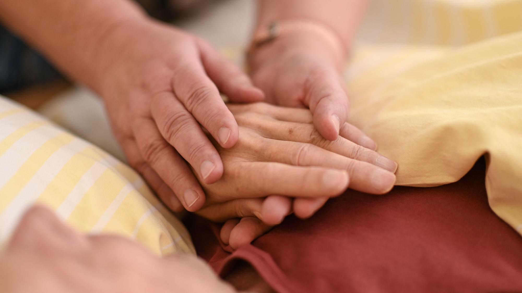 Eine Person hält mit beiden Händen die Hand eines Menschen, der mit einer gelben Decke zugedeckt ist.