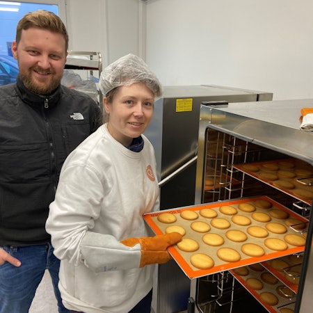 Kerstin Hottewitzsch und Nicolas Gritto stehen vor einem Industrie-Ofen. Hottewitzsch hält ein Blech mit Keksen und schiebt es in den Ofen. Sie lächeln beide in die Kamera.