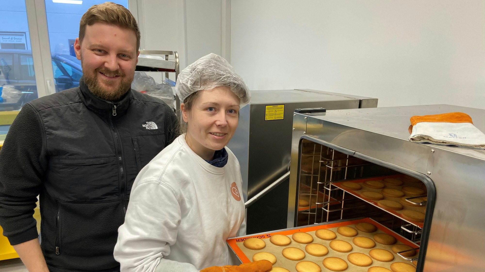 Kerstin Hottewitzsch und Nicolas Gritto stehen vor einem Industrie-Ofen. Hottewitzsch hält ein Blech mit Keksen und schiebt es in den Ofen. Sie lächeln beide in die Kamera.