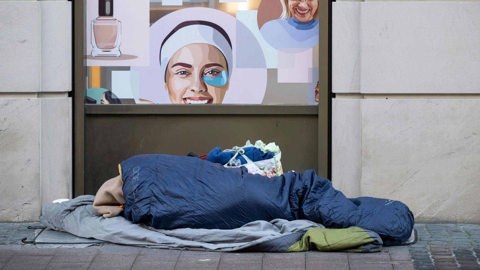 Das Lager eines wohnungslosen Menschen vor einer Hauswand unterhalb einer Kosmetik-Werbung.